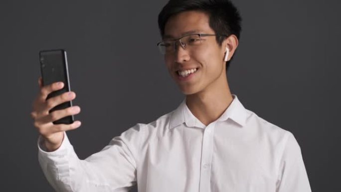 年轻英俊的亚洲男子戴眼镜快乐地在灰色背景下用手机视频聊天