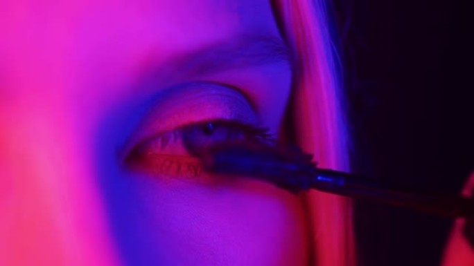 霓虹灯下的眼妆。一个女人用睫毛膏画眼睛，她的脸被彩灯照亮。时尚和美丽。