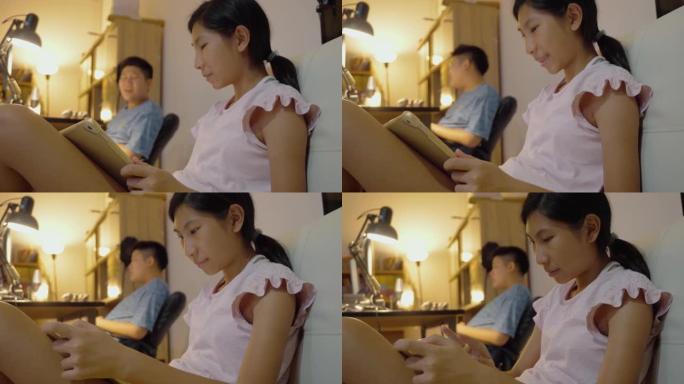 亚洲女孩用数字平板电脑看电影在她的哥哥附近，她的哥哥在家里晚上用电脑玩网络游戏，生活方式概念。