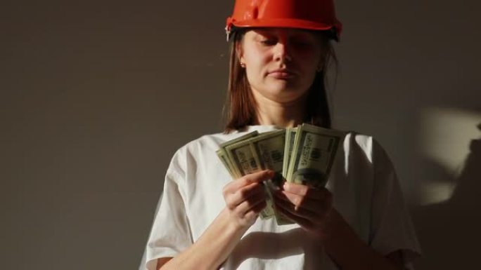 年轻的微笑女工戴着橙色安全帽数钱，站在公寓的建筑工地。建筑师持有现金用于房屋处理