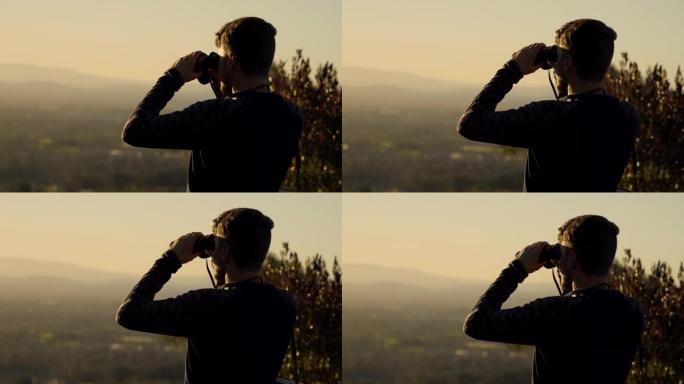 男子站在山顶的了望，用双筒望远镜观看。