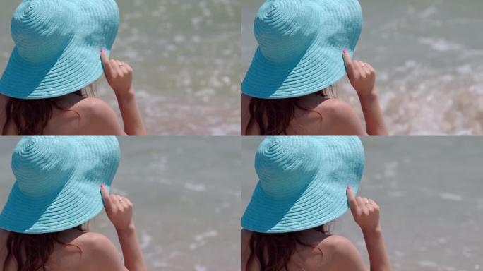 戴着蓝色帽子的女人在海滩享受日光浴。在海边享受微风的晒黑女人的特写脸。背景中的海洋。慢动作4k