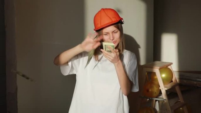 年轻的微笑女工戴着橙色安全帽数钱，站在公寓的建筑工地。建筑师在办公室里扔和玩美元钞票