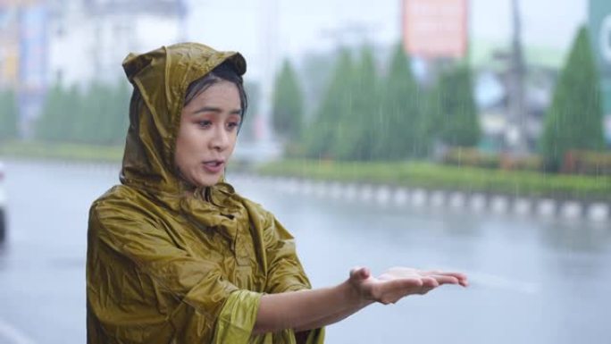 年轻疲惫的女上班族穿着黄色雨衣，被雨水卡住，站在屋顶下，用手玩耍收集雨滴，季风造成的大雨，亚洲城市生