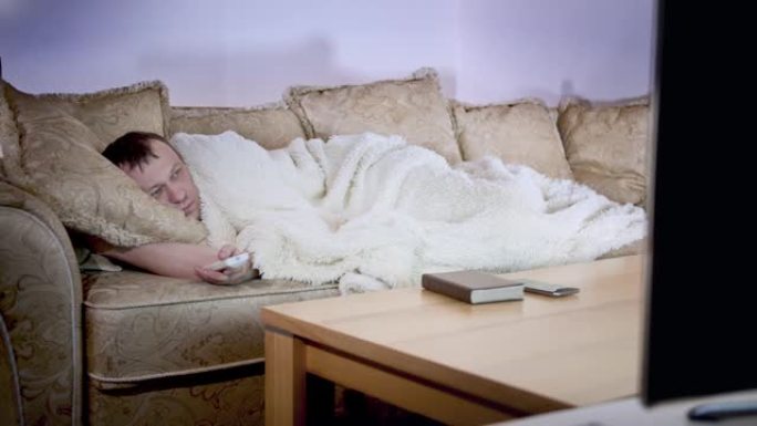 一名男子躺在沙发上盖着毯子，切换电视频道，晚上照明