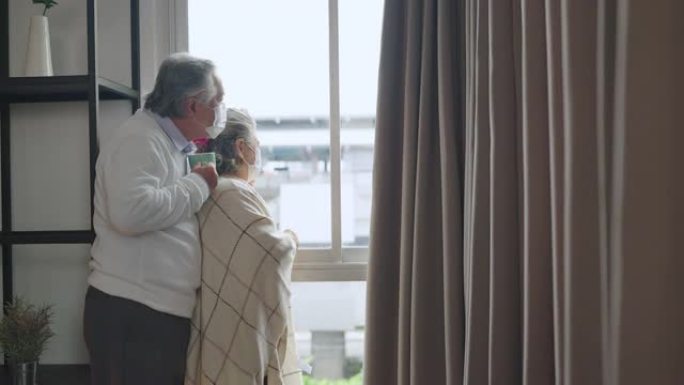 亚洲退休的老年夫妇戴着口罩透过他们的家庭窗户看着世界，拥抱和支持在一起，亚洲人的家庭隔离社会距离保持