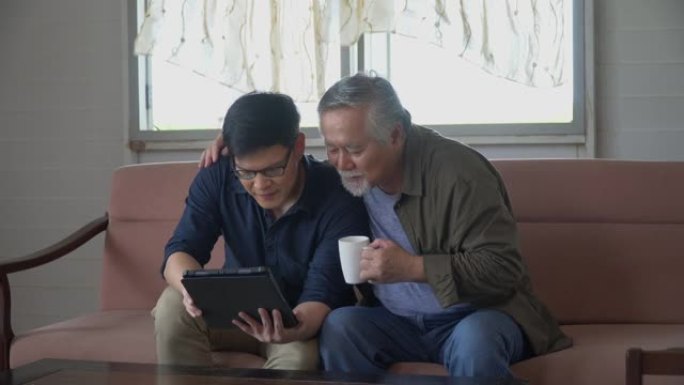 快乐的亚洲小儿子和资深老父亲坐在沙发上一起使用平板电脑在家