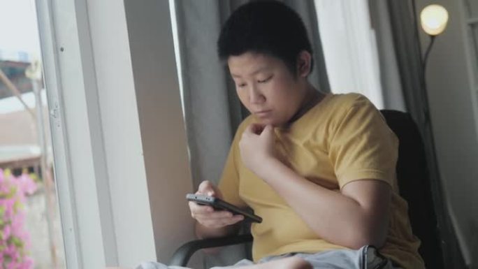 亚洲男孩坐在家里的窗户旁，通过智能手机观看电影或病毒视频，这是生活方式的概念。