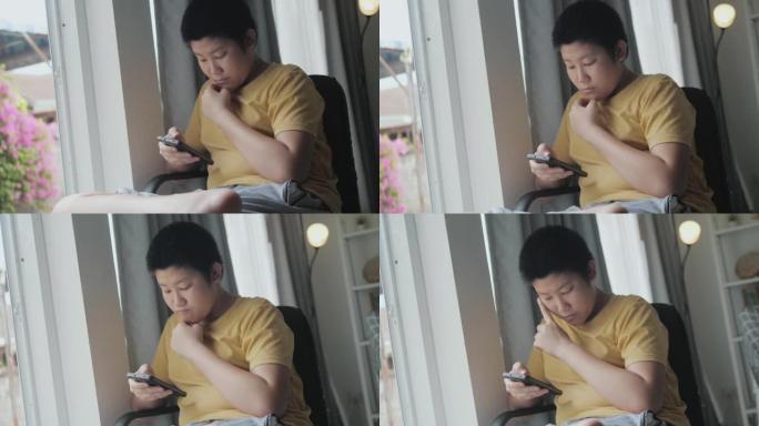 亚洲男孩坐在家里的窗户旁，通过智能手机观看电影或病毒视频，这是生活方式的概念。