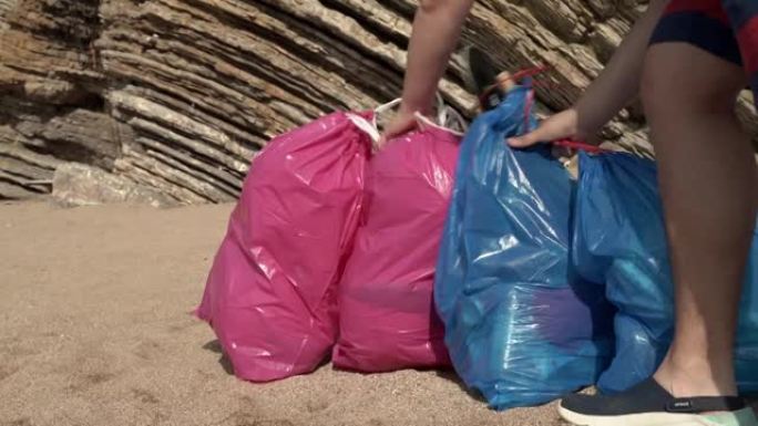 男子志愿者在阳光明媚的夏日带着装满垃圾的大彩色袋子从沙滩上带走