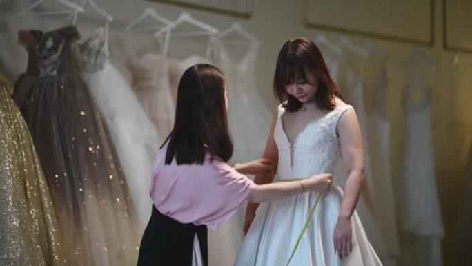 亚洲中国女性婚纱店老板为定制设计婚纱测量她的顾客新娘身材