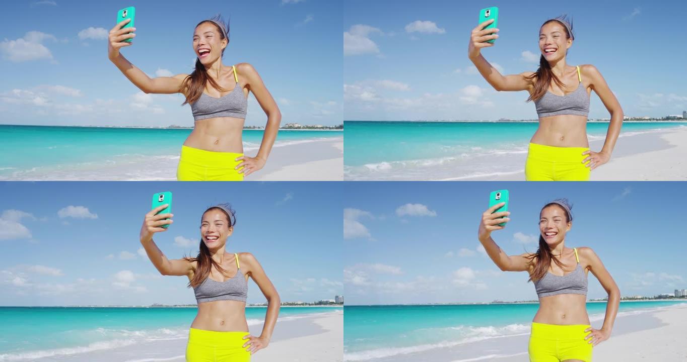 运动清新的女人在沙滩上跑步后笑着用手机自拍。女孩在暑假锻炼后进行健身自拍，过着健康的生活方式。红色慢