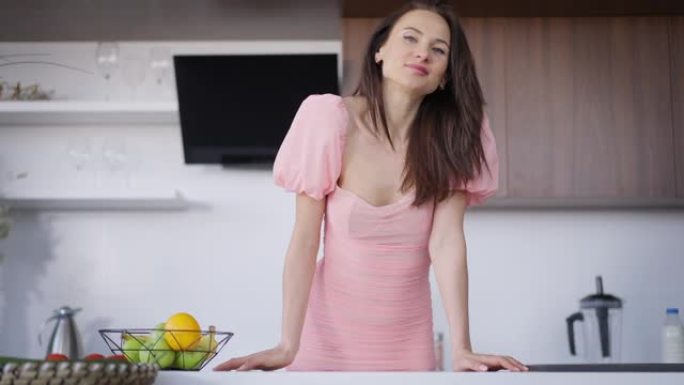 年轻苗条美丽的黑发女人站在厨房台面上微笑着看着相机的照片。积极自信迷人的粉红色连衣裙女士在家摆姿势