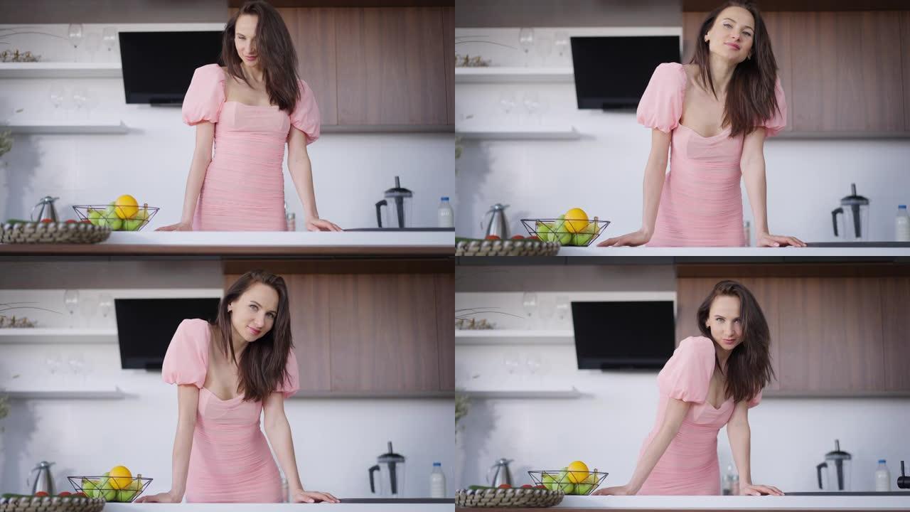 年轻苗条美丽的黑发女人站在厨房台面上微笑着看着相机的照片。积极自信迷人的粉红色连衣裙女士在家摆姿势