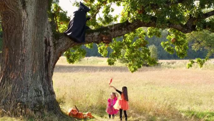 两个女孩站在一棵巨大的树下，一棵穿着黑色雨衣的收割者坐在上面，拿着一个大糖果，用糖果戏弄孩子们