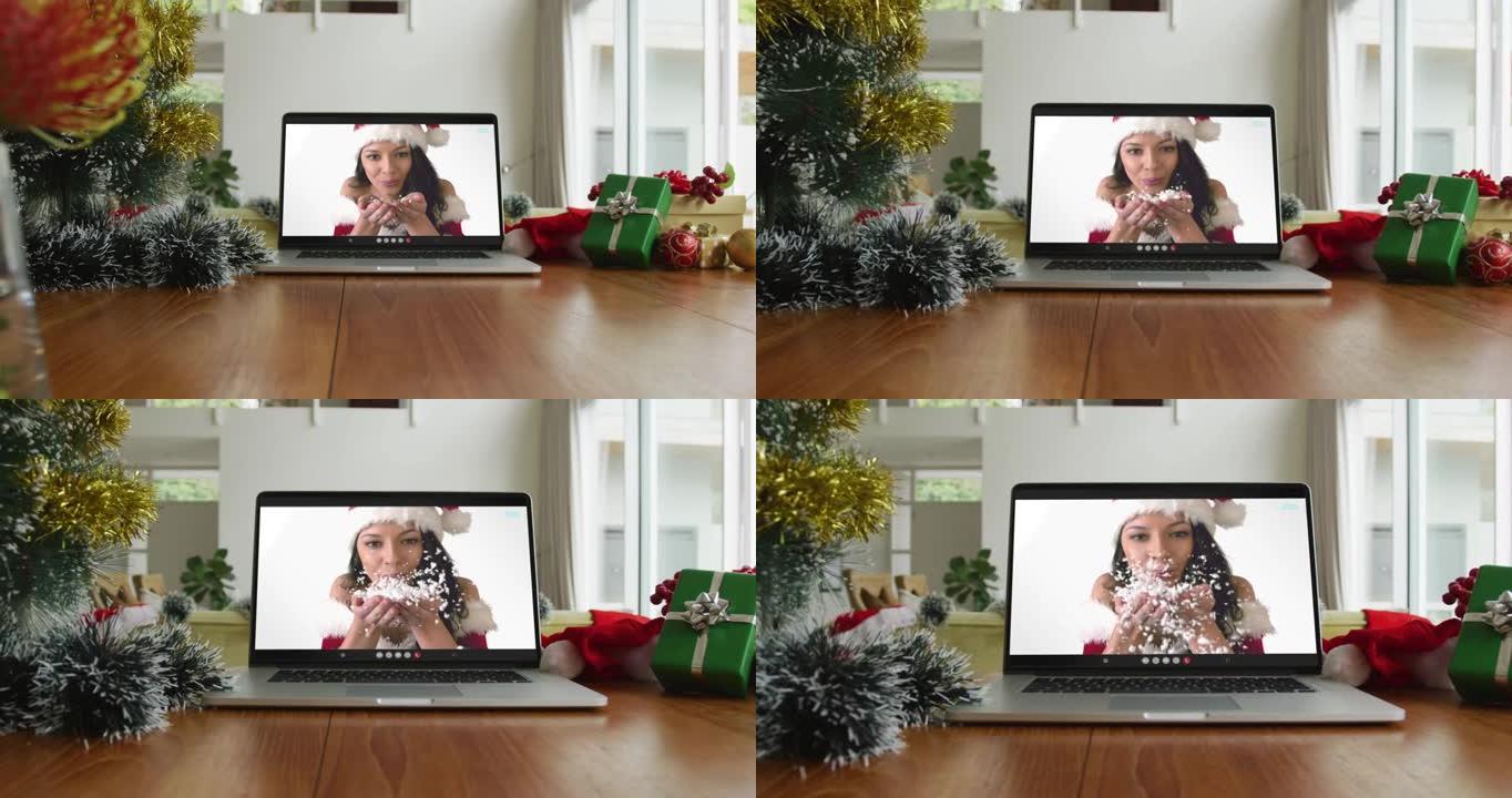 微笑的白人妇女戴着圣诞老人的帽子，在笔记本电脑上的圣诞节视频通话中吹雪