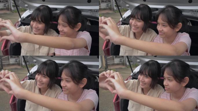 两个亚洲可爱的女性青少年坐在掀背车的后侧，喜欢一起做手势心形。年轻的生活方式。
