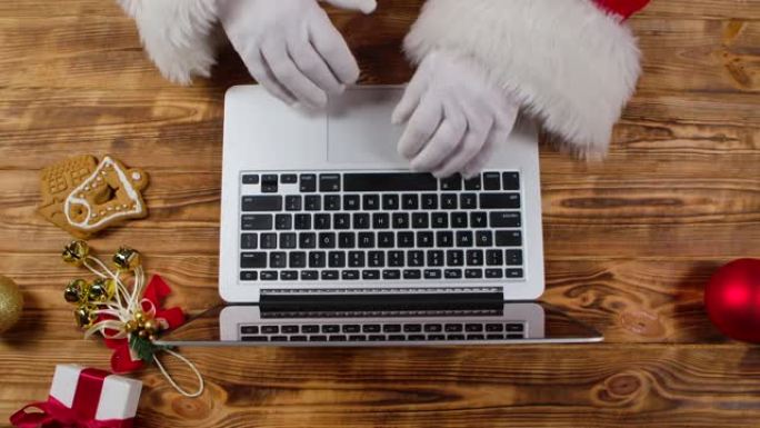 顶视图圣诞老人双手戴着白手套在木制新年装饰桌子上的键盘上打字。圣诞老人与笔记本电脑一起工作，浏览邮件