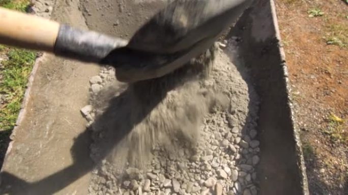 用铲子将水泥倒入带有小岩石的沙子上，以形成混凝土混合物