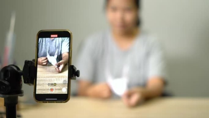 亚洲妇女，年轻的成年人在家里的智能手机上在线制作纸鸟折纸拍打鸟。冠状病毒封锁检疫期间新的正常生活方式