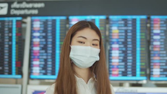 年轻的亚洲妇女站在机场出发板前戴着口罩看相机