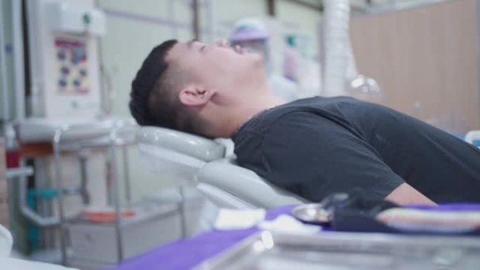 特写一名亚洲男性患者在电动牙科椅上缓慢水平躺下，年度口腔治疗准备程序，背景模糊的外科员工站在关心