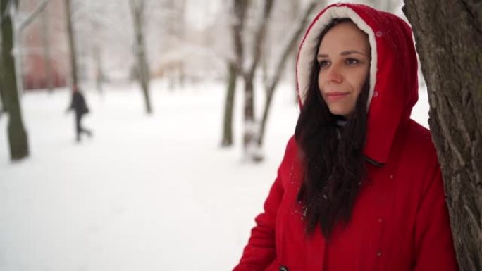 冬季穿着红色夹克和兜帽的年轻女子的肖像。美丽的女士站在树旁，移开了目光。蓬松的雪包围了周围的一切
