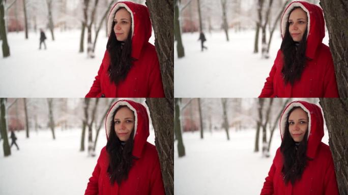 冬季穿着红色夹克和兜帽的年轻女子的肖像。美丽的女士站在树旁，移开了目光。蓬松的雪包围了周围的一切