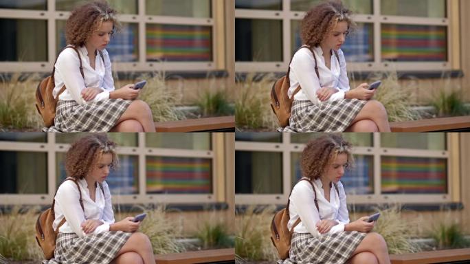 可爱的十几岁的女学生坐在学校大楼附近的长凳上，手里拿着智能手机