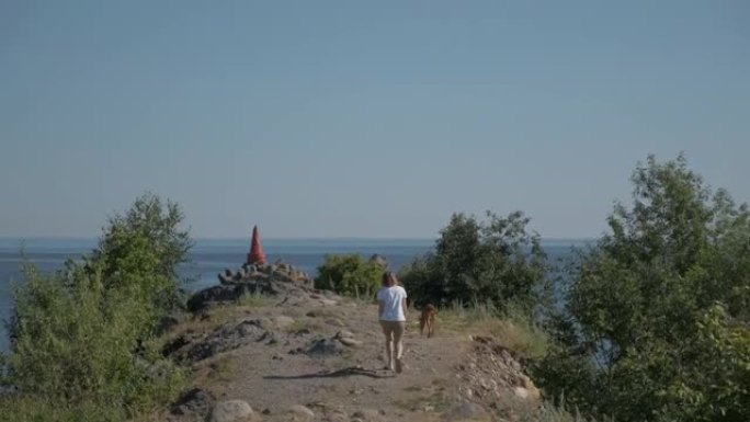 一名年轻女子带着法老品种的红狗沿着海岸旅行。夏天阳光明媚的日子，蓝天。