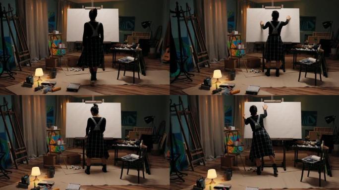 黑暗艺术工作室的女人的影子走近一个大画架，用白色画布围绕着乱七八糟的油漆，画笔，女孩触摸结构，检查画