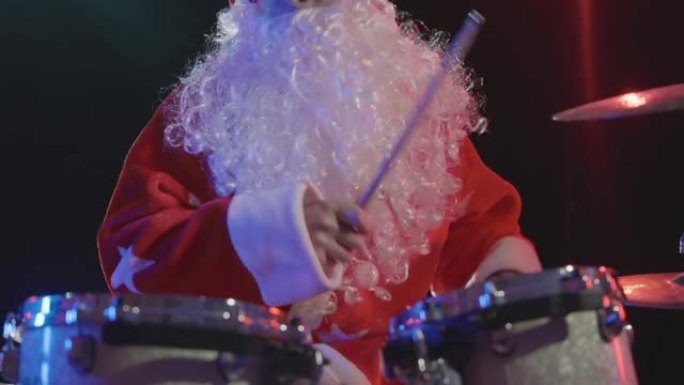 圣诞老人的节日表演。一个穿着新年服装，留着胡须的男人现场演奏鼓包，用棍棒积极敲击鼓和敲击钹。特写。慢