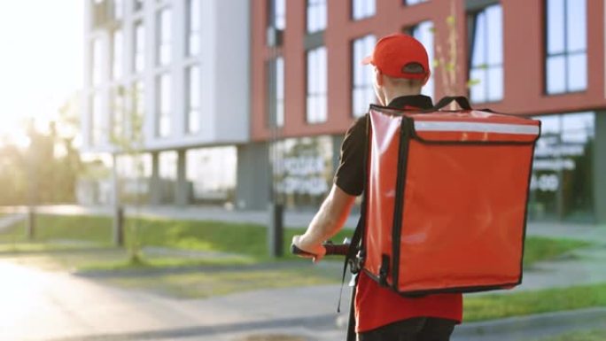 交付男性包裹递送服务人员骑着电动滑板车在日落时带着红色背包在街上的空人行道上。无法识别的年轻快递员骑
