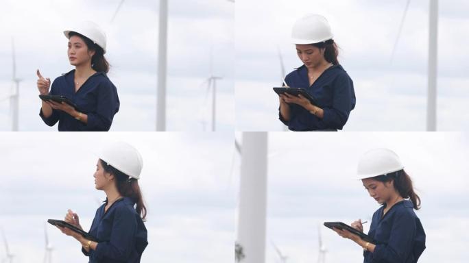 亚洲女性工程师在风力涡轮机农场工作，手持平板电脑和笔记本电脑，她戴着白色安全帽