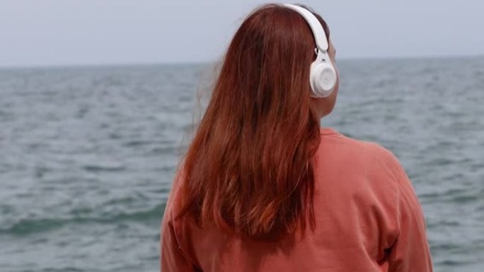 年轻女孩的特写肖像在海滩上看着大海，戴着无线耳机在户外冥想
