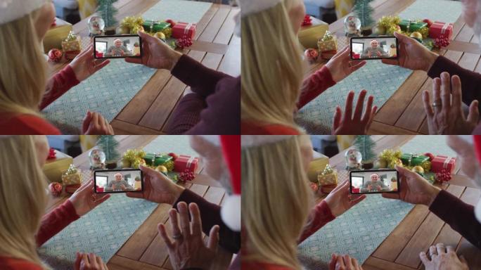 白人夫妇戴着圣诞老人帽子，使用智能手机与屏幕上的男人进行圣诞节视频通话