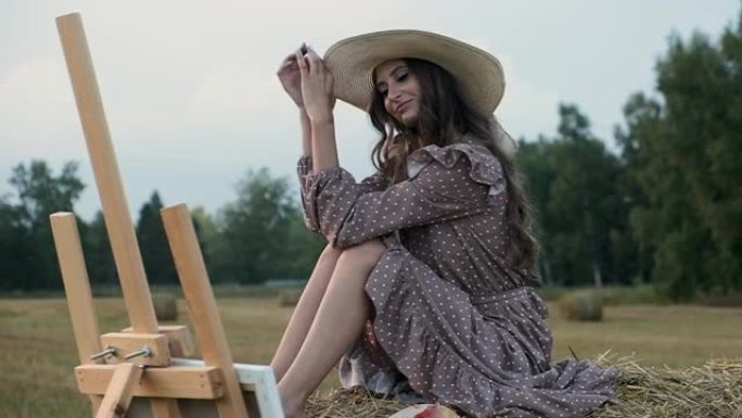 穿着太阳裙和帽子的幸福女人坐在干草堆上，看着彩绘的照片