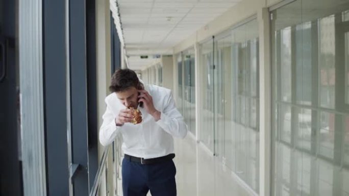 年轻的办公室经理穿着西装吃汉堡，并在智能手机上接听电话