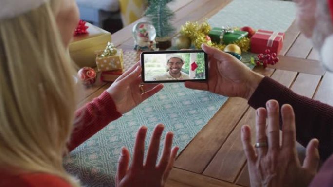 白人夫妇戴着圣诞老人帽子，使用智能手机与屏幕上的朋友进行圣诞节视频通话