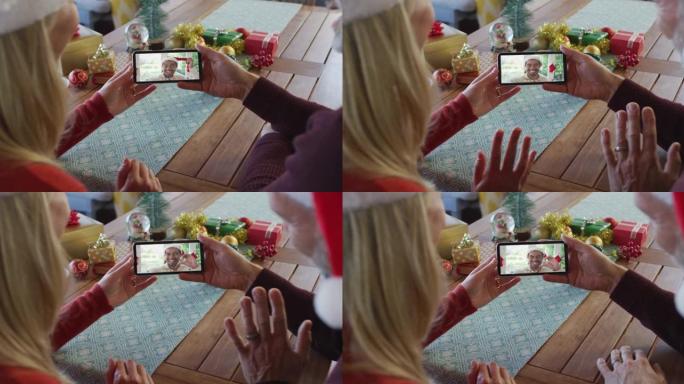 白人夫妇戴着圣诞老人帽子，使用智能手机与屏幕上的朋友进行圣诞节视频通话