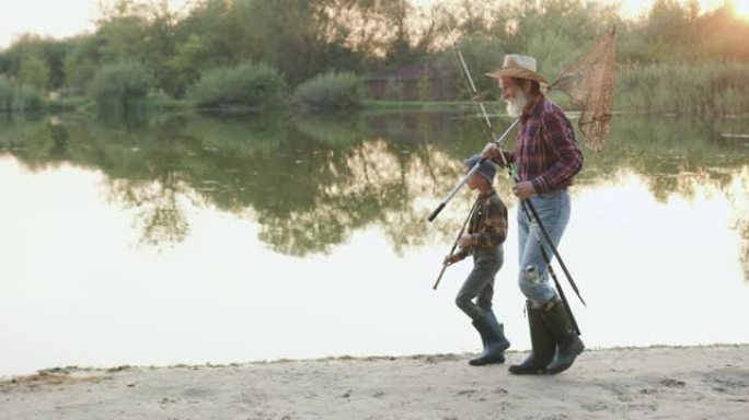 好看的积极友好的成熟的大胡子菲什曼和他好奇的小孙子一起背着钓具，在池塘的岸边钓鱼