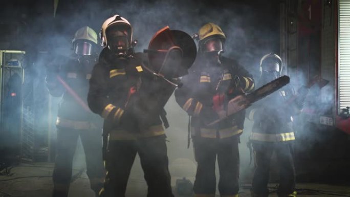 一组消防员戴着全套设备，氧气面罩，紧急救援工具，圆形液压和气锯，斧头和大锤。背景中的烟雾和消防车。