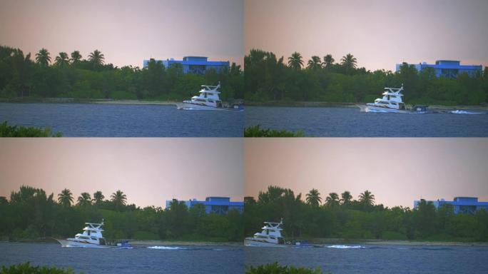 一艘白色渔船穿过泻湖。船从右向左移动。在背景中，海岸密布，棕榈林。粉红色的夜空。