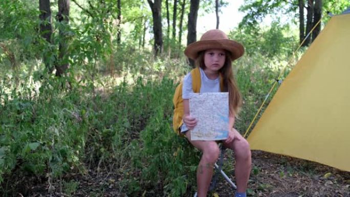 戴着帽子，背着背包的小女孩若有所思地坐在帐篷附近的森林里。夏令营里的孩子手里拿着地图。儿童地图阅读技