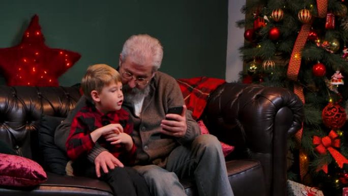 优雅的老人和小男孩在智能手机上浏览内容，玩得开心。祖父和孙子坐在靠近发光圣诞树的装饰房间的沙发上。特