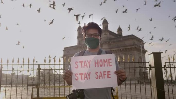 一个戴着口罩的年轻人站着，举着标语牌，上面写着 “待在家里保持安全”。