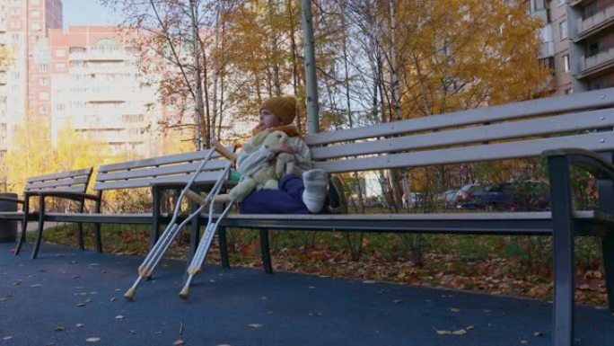 孩子拄着拐杖在秋季公园散步。坐在公园长椅上玩玩具的孩子。女孩的一条腿骨折了。