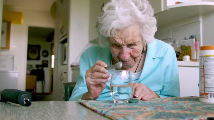 老妇人仔细地看着，同时小心地将1/4茶匙的橙汁混合饮料倒入水中，作为每日维生素补充剂或药物