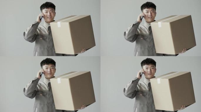这位年轻的亚洲快递员穿着制服，手里拿着一个大购物盒，牛皮纸纸箱和物流给客户打电话