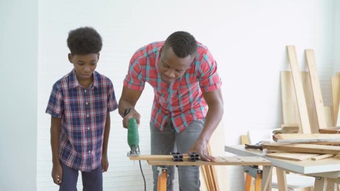 好奇的非洲裔美国儿童男孩在工厂里看着并学习父亲用电锯切割一块木头